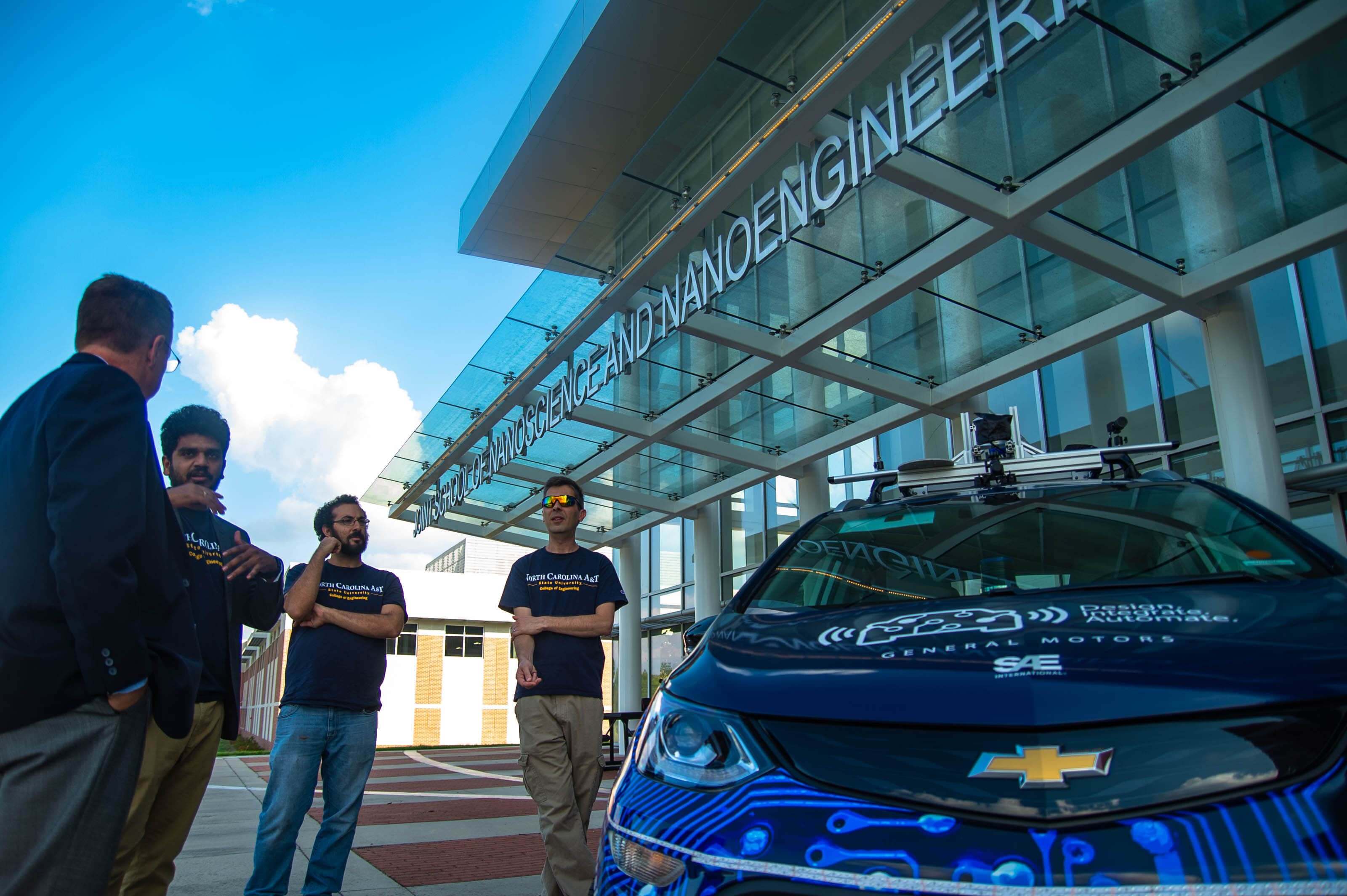 Four people stand outside a nanoscience and nanoengineering building, next to a car carrying equipment on its roof.