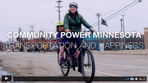 Person riding a bicycle in the roadway of an urban setting with a child on the back, smiling. Text on screen: Community Power Minnesota - Transportation and Happiness