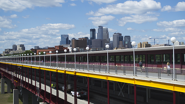 City skyline of Minneapolis rising in the background above a foreground view of Washington Avenue Bridge on UMN campus