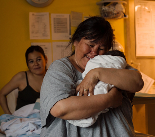 Smiling Indigenous doula holds baby while birth person looks on