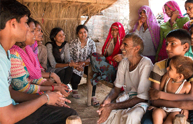 A multi-generational group sits happily under a a grass shelter