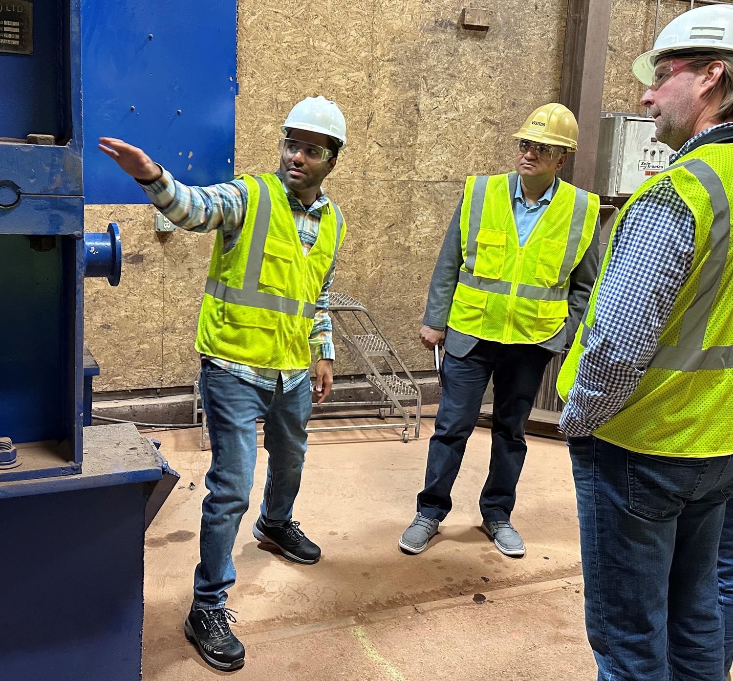 Three people stand around industrial equipment with protective vests/hats. One person is gesturing towards equipment.