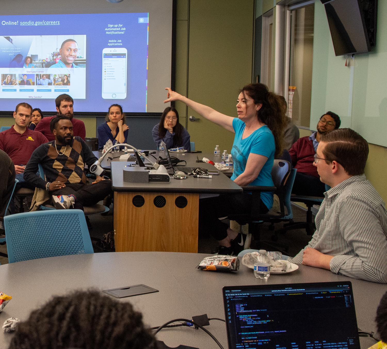 Person stands in a classroom, gesturing to a powerpoint screen