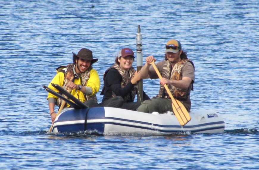 Researchers paddle an inflatable raft on a lake, with one holding a sediment core.