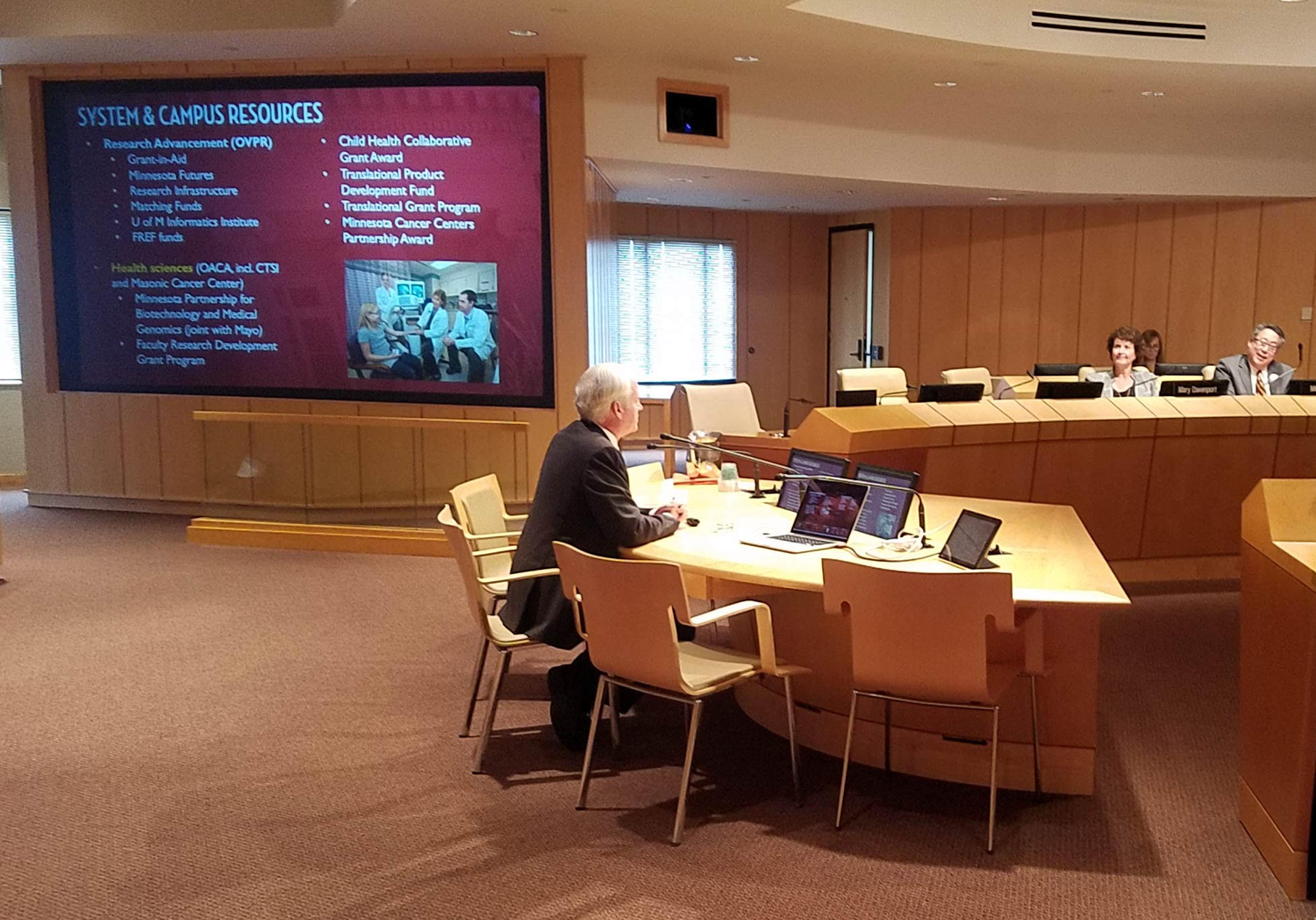 VPR Cramer sits at a table to present at the October 11 Board of Regents meeting. Slide from his presentation is projected behind him.