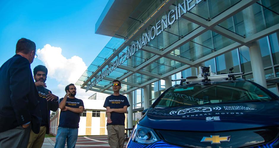 Four people stand outside a nanoscience and nanoengineering building, next to a car carrying equipment on its roof.