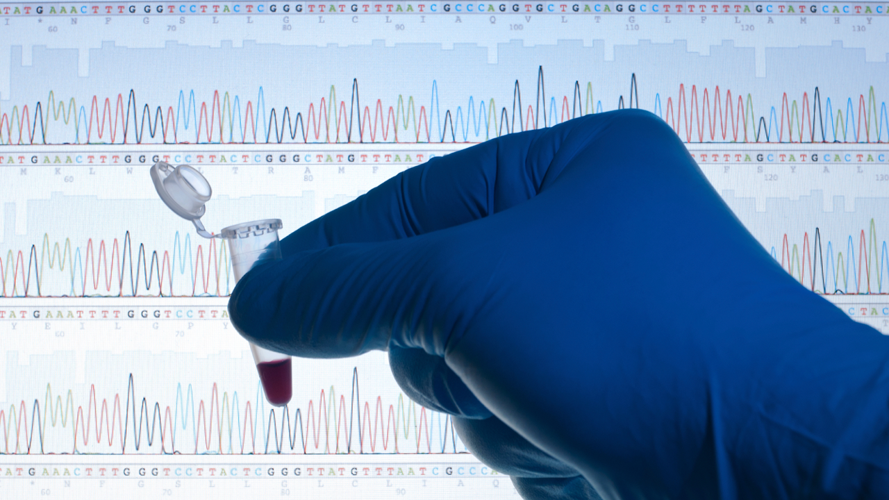 A gloved hand holds a vial in front of a DNA sequence on a screen